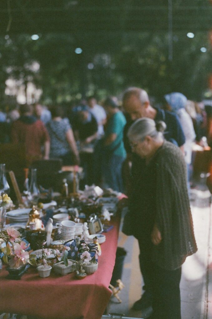 A busy outdoor market with people examining various antique items on a table.